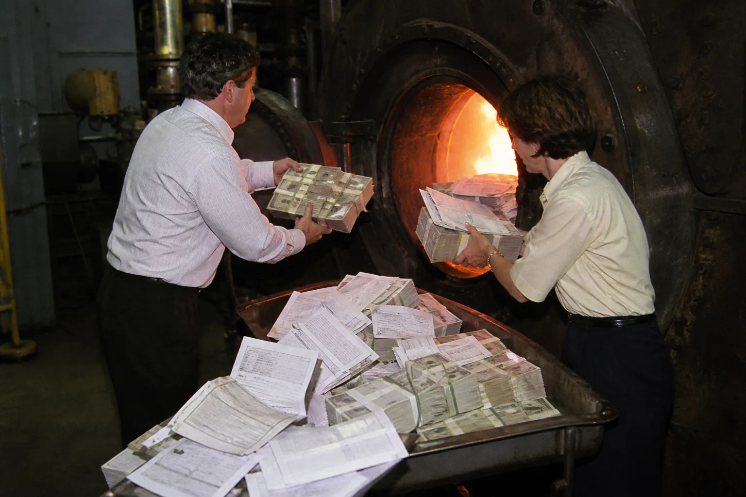 Two people feed stacks of cash into an industrial furnace.