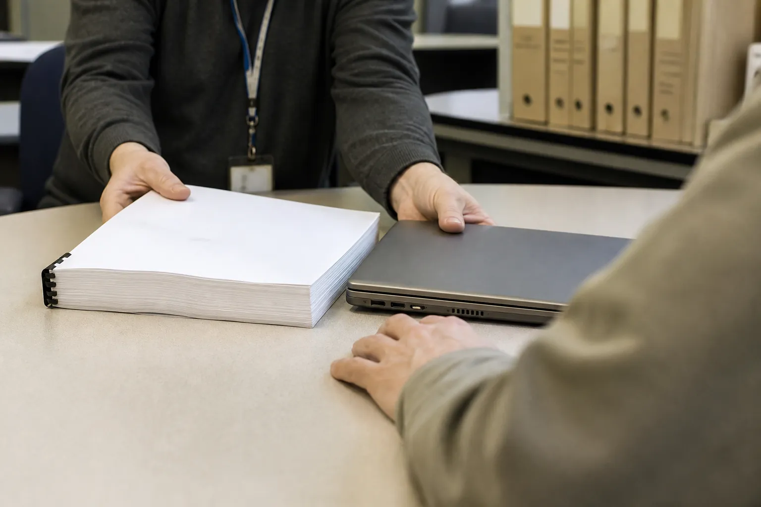 Hands sliding a thick printed report across a desk toward a laptop.